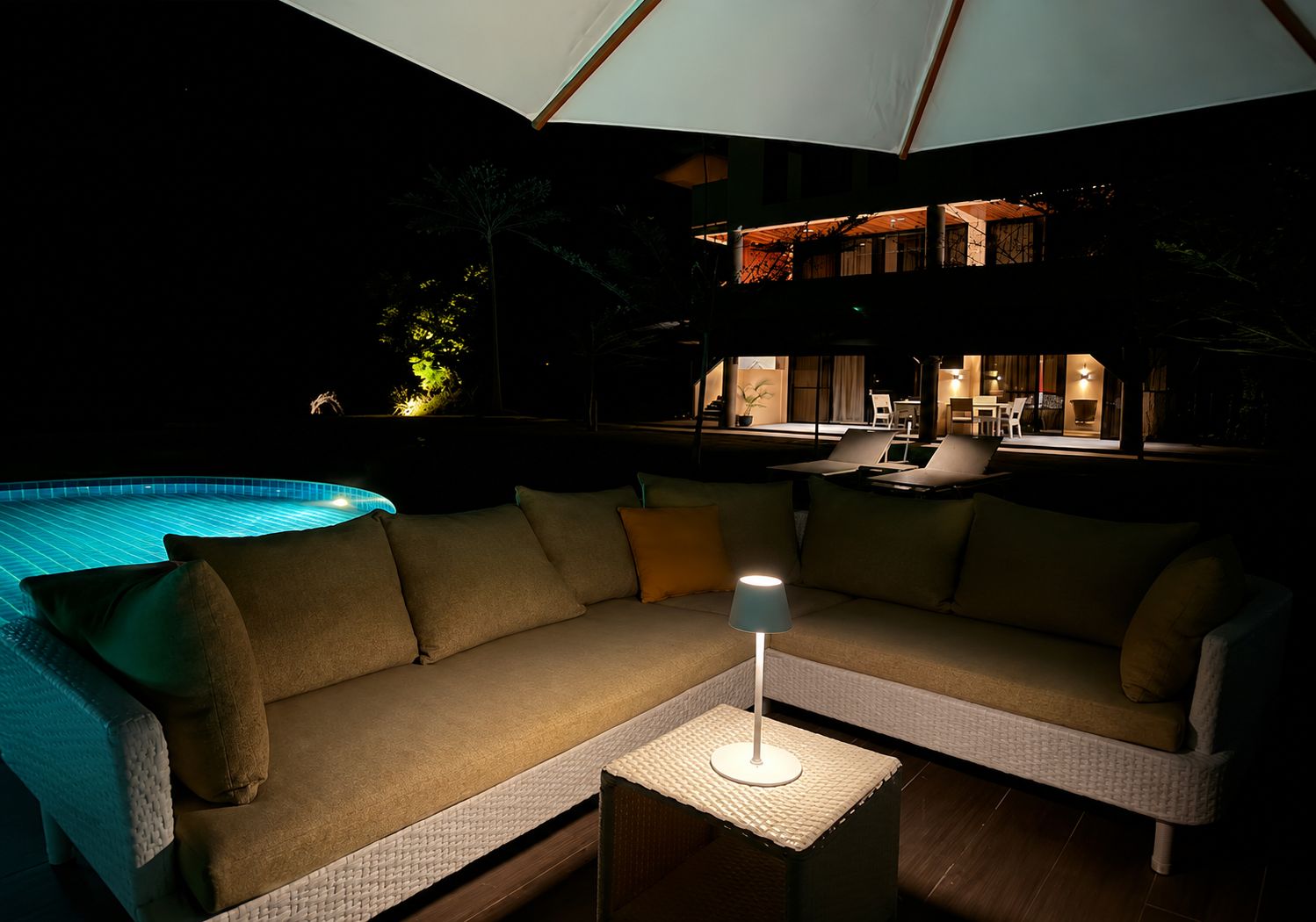 Un espacio al aire libre con mesas y sillones para disfrutar de la noche, una piscina y una lámpara sobre una mesa.