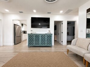 A modern living room with a flat-screen TV mounted on the wall, a teal cabinet, a beige sofa, and a wooden floor. - A First-Class Stay in Tampa Bay | Stadium, Midtown, and Downtown (Tampa)