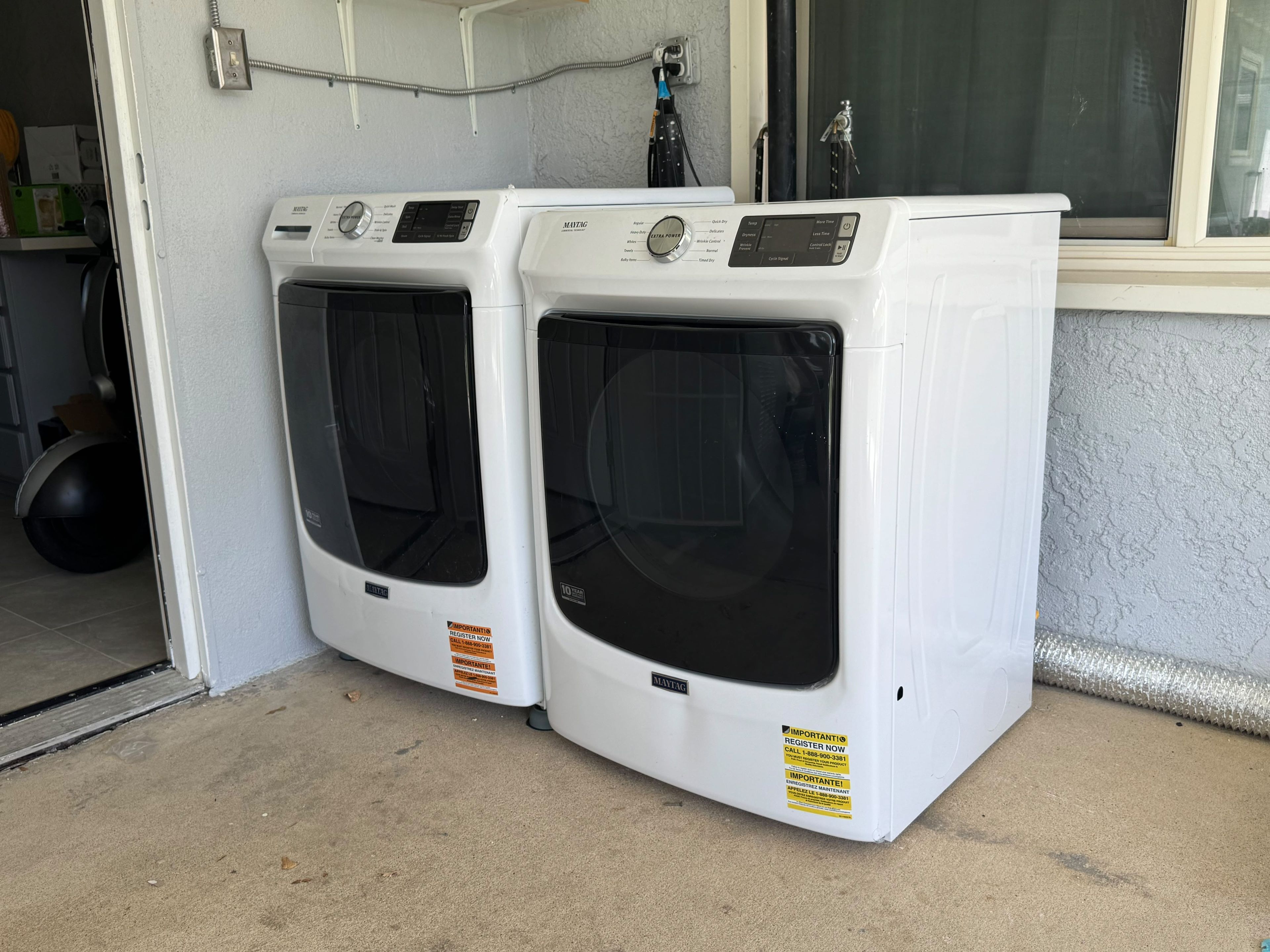 Two white front-loading washing machines with control panels, placed next to a window with a metal frame and a water spout.
