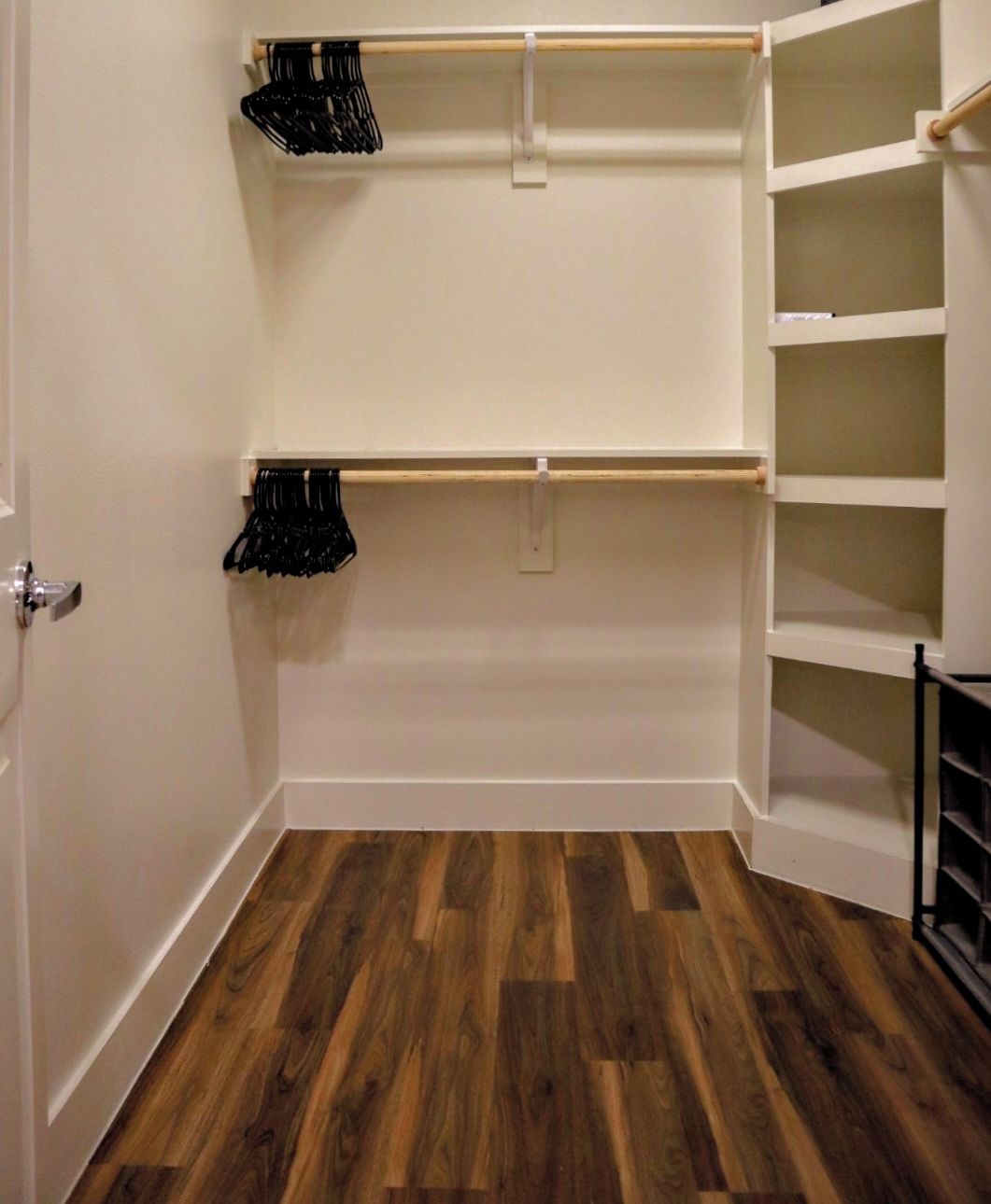 Un armario con repisas abiertas y espacio para colgar, piso de madera y una puerta con picaporte plateado.