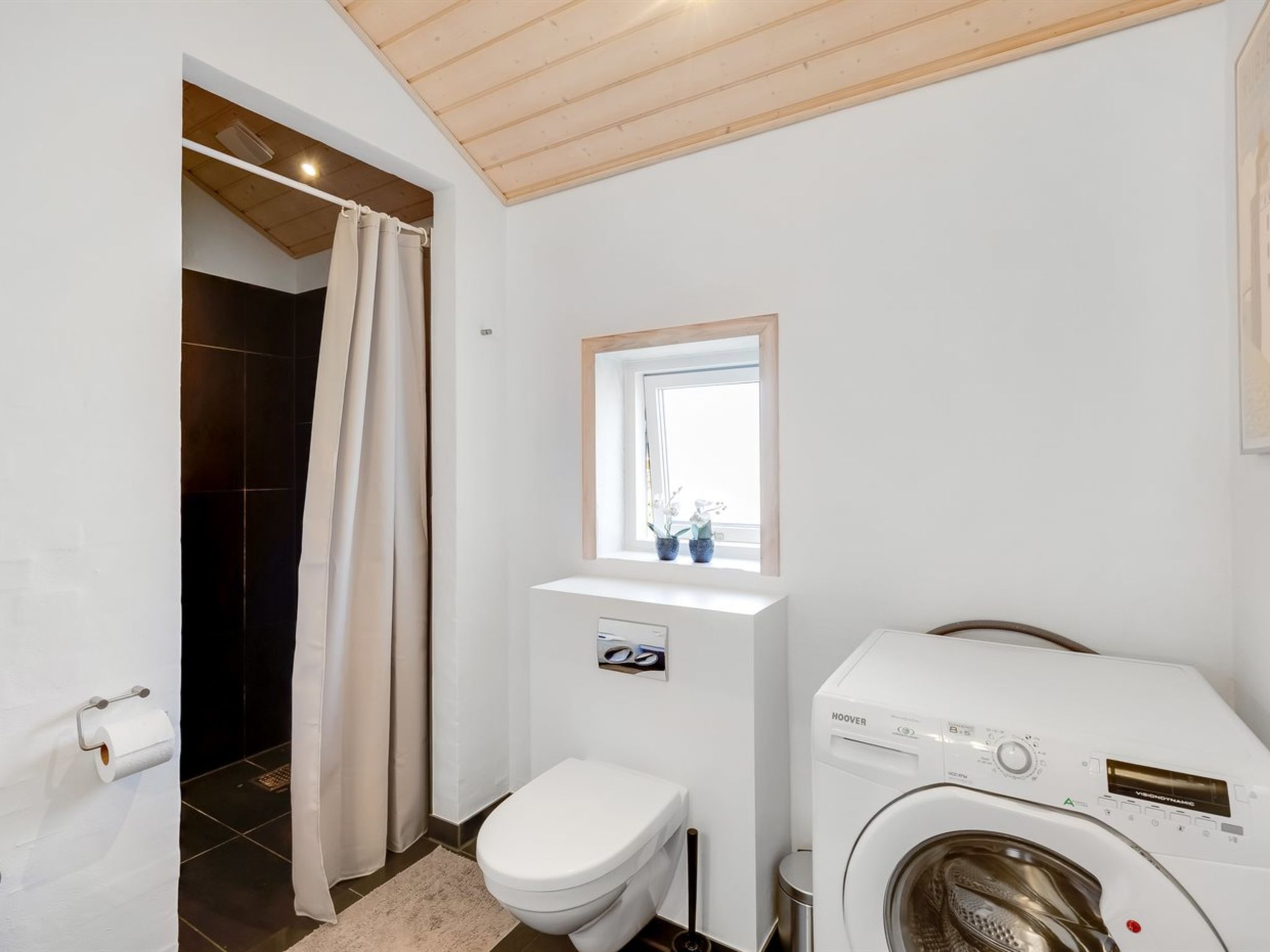 A compact bathroom with a toilet, a washing machine, and a shower area.