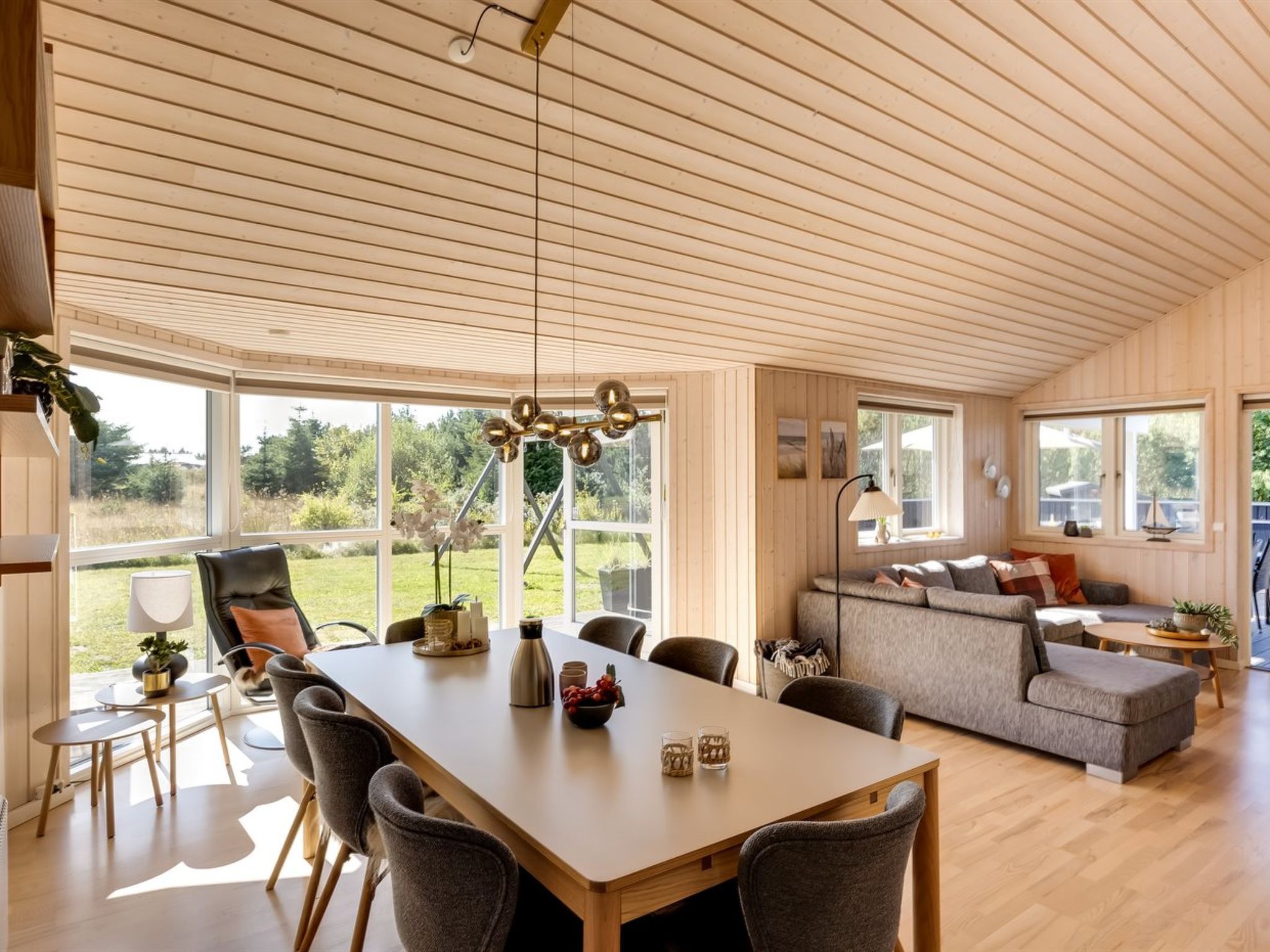 A spacious living area with a wooden ceiling, a dining table with chairs, a sofa, and a large window offering a view of greenery.