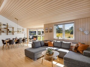 Una sala de estar moderna con techo de madera, un sofá seccional gris, una zona de comedor con mesa y sillas de madera, y un ventanal con vistas a zonas verdes.