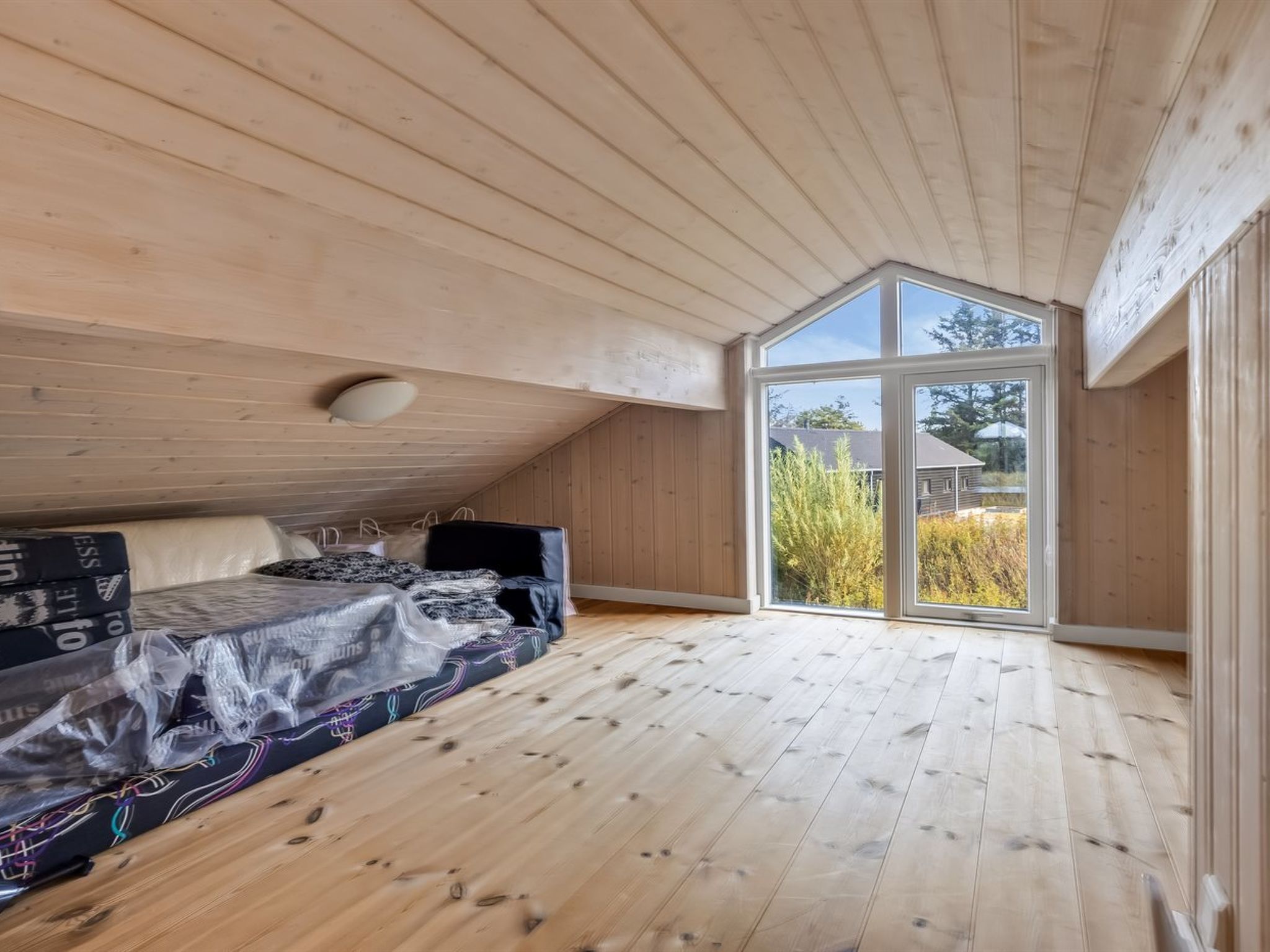Une pièce avec un plafond en pente, un plancher en bois, un lit recouvert d’une couvre-lit à motifs et une grande fenêtre donnant sur un paysage verdoyant et un bâtiment situé à l’extérieur.