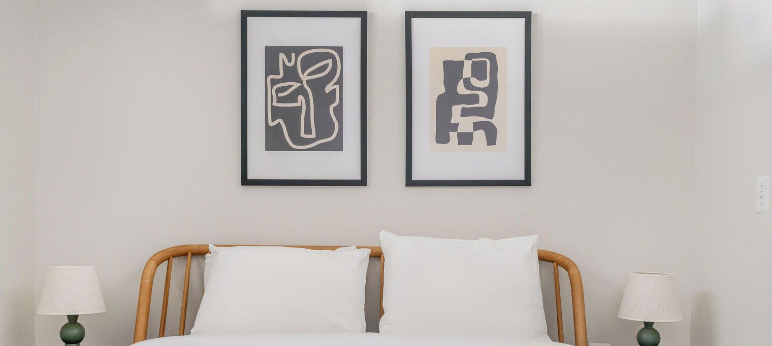 Una cama con ropa blanca, dos cuadros abstractos enmarcados encima y dos lámparas de noche a cada lado.