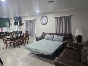 Un salon avec un canapé, une table à manger et des chaises, une horloge au mur et une lampe sur une table d’appoint.