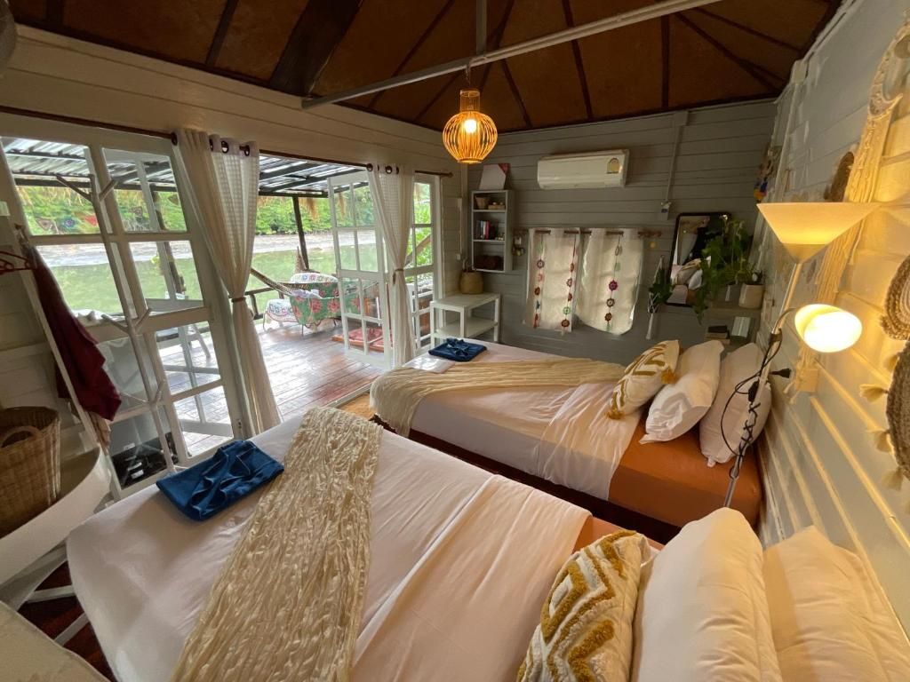 A room with two beds, a table, and a chair. There is a window with curtains, a hanging light fixture, and a wall-mounted air conditioning unit.