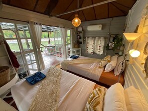 Habitación con dos camas, una mesa y una silla. Se ve una ventana con cortinas, una lámpara colgante y un aire acondicionado instalado en la pared.