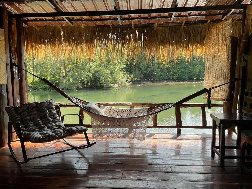 A hammock chair and a table on a wooden deck with a view of a lake and forest.