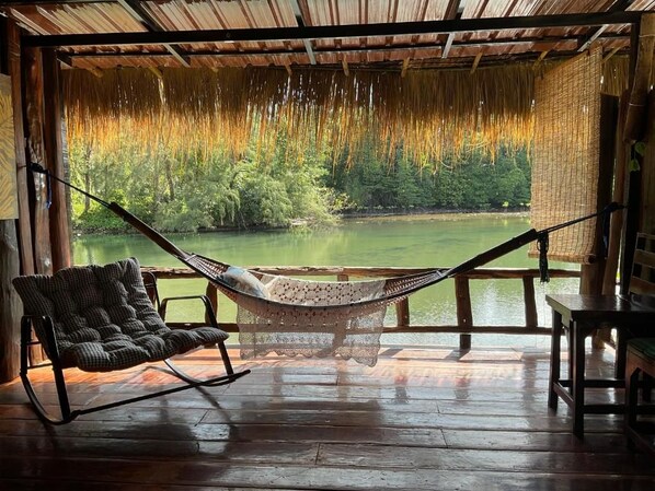 Una silla hamaca y una mesa en una terraza de madera con vista a un lago y un bosque.