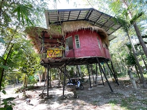 A red wooden house with a thatched roof, elevated on stilts, surrounded by trees.