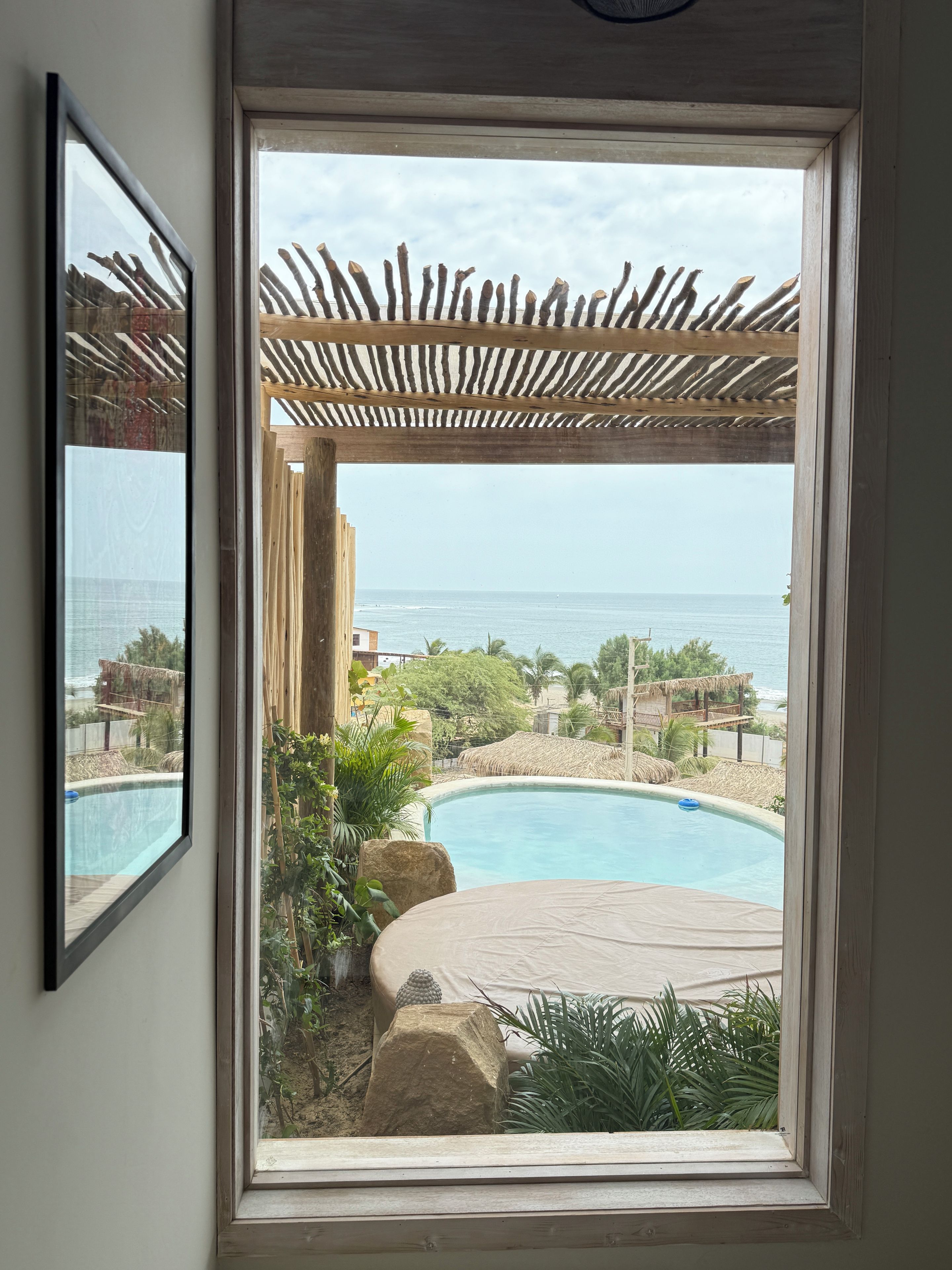 A window view of a pool area with a wooden pergola, a clear sky, and a distant view of the sea.