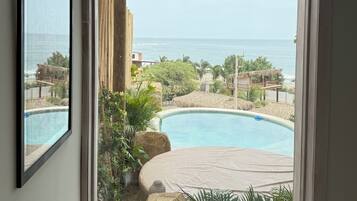 A window view of a pool area with a wooden pergola, a clear sky, and a distant view of the sea.