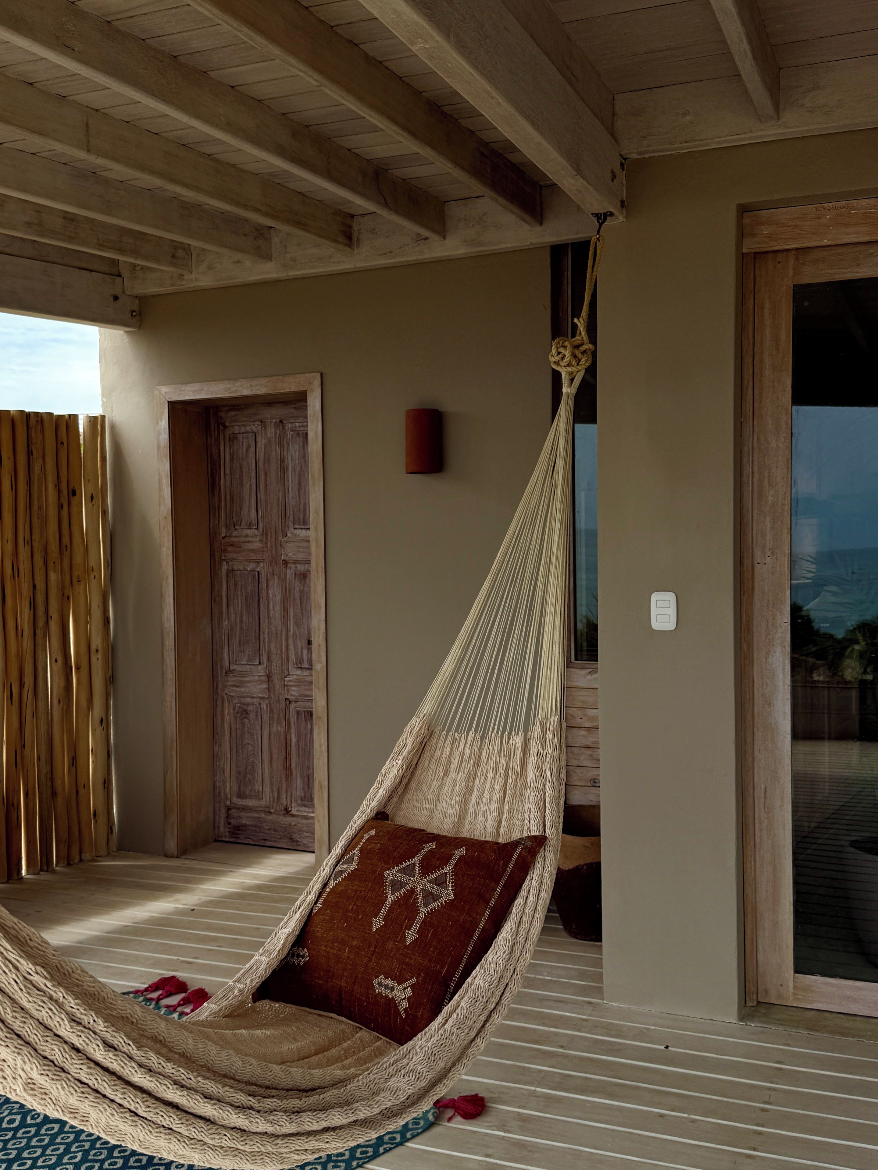 Una silla hamaca con un cojín estampado sobre una terraza de madera, una puerta de madera y vistas a través de un ventanal.