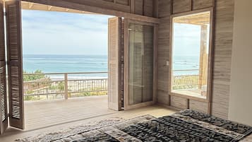 Een slaapkamer met een groot bed, houten balken in het plafond en uitzicht op zee door open deuren.