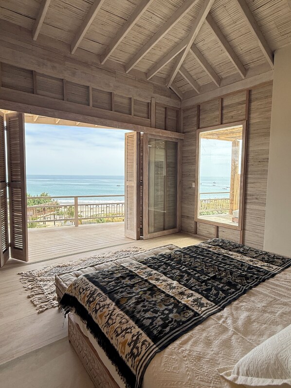 A bedroom with a large bed, wooden ceiling beams, and a view of the ocean through open doors.