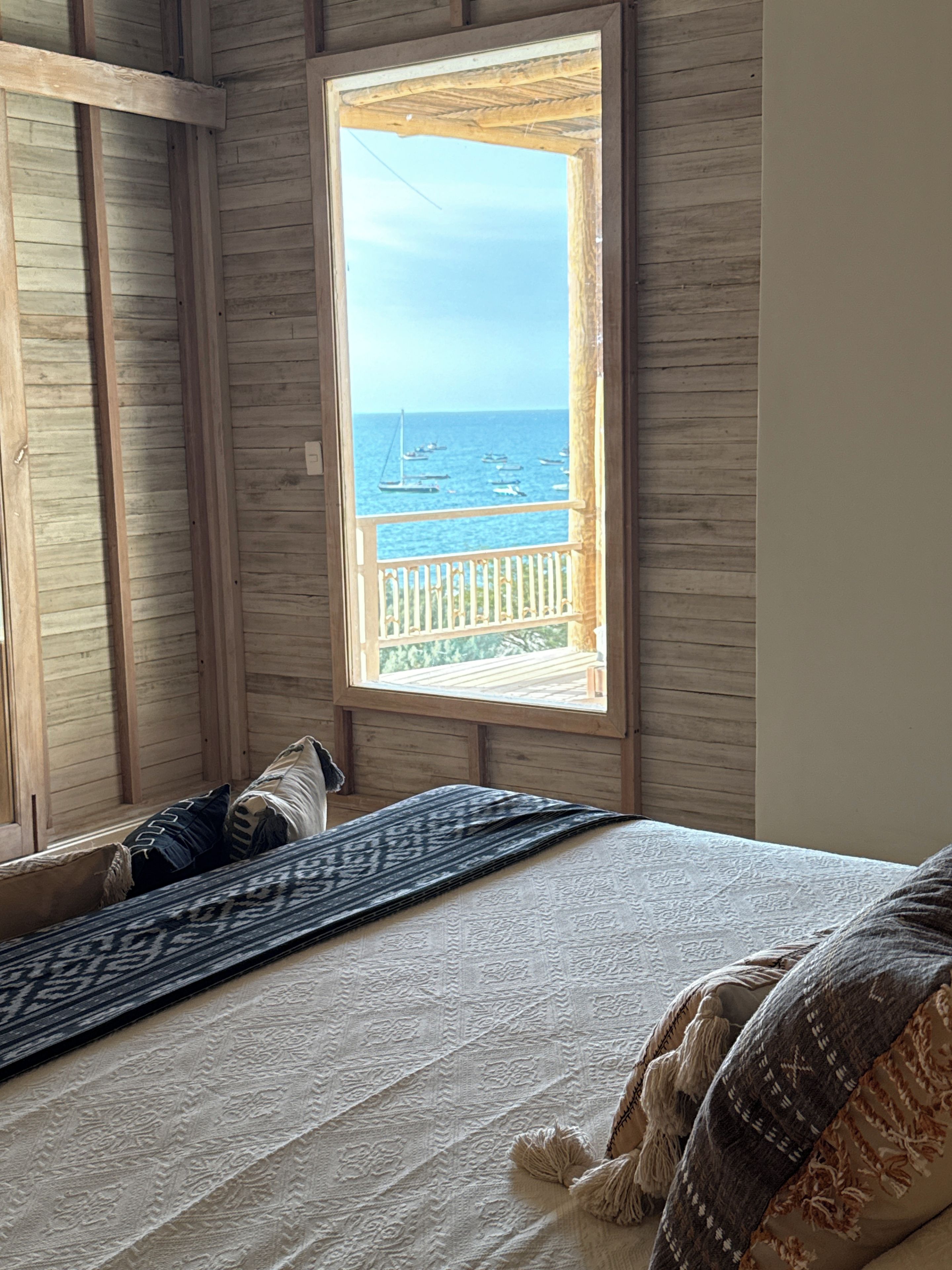 A room with a bed, a window with a view of the sea, and wooden walls.