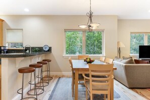 A modern kitchen with a dining area, bar stools, and a large window offering a view of greenery. - NEW 2BR w/ Hot Tub, Free Shuttle, Parking, WiFi (Vail)
