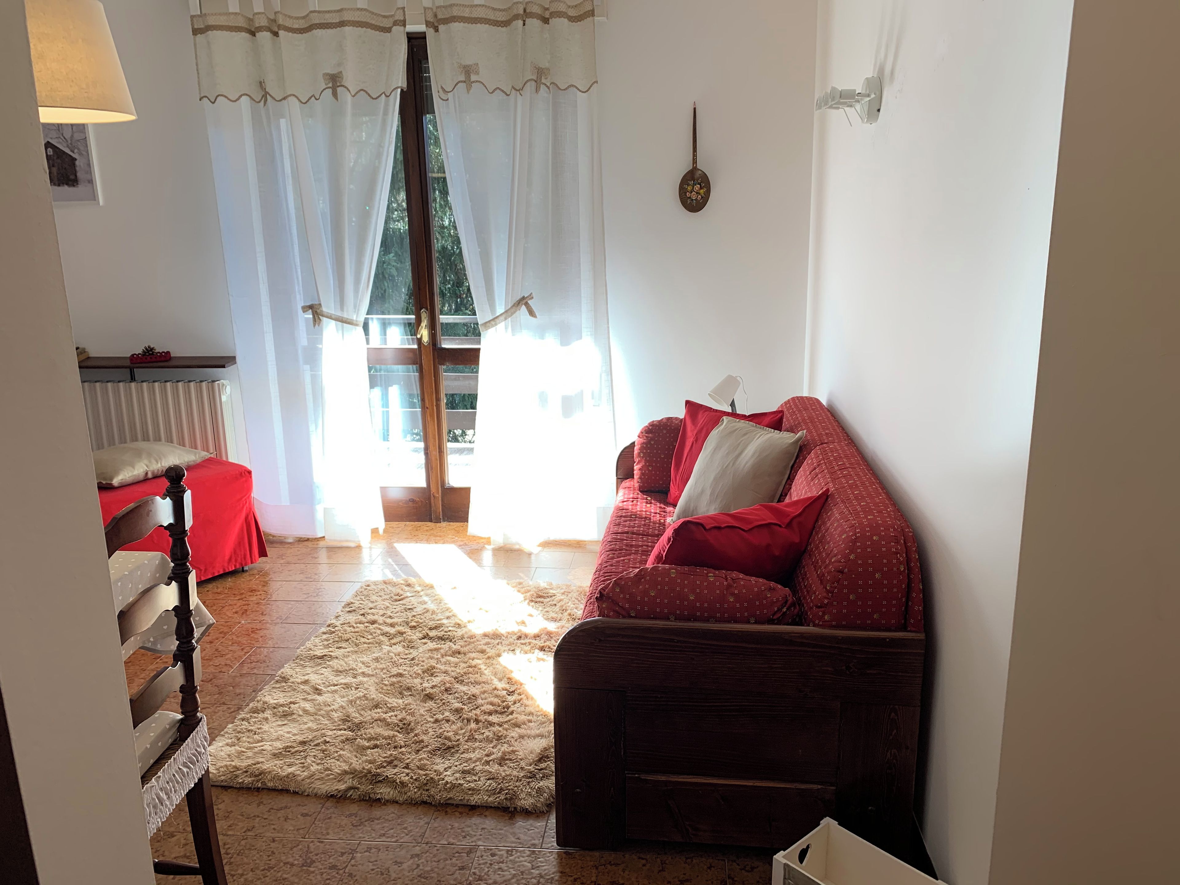 Una sala de estar acogedora con un sofá rojo, una silla de madera, un tapete mullido y vistas a través de una ventana con cortinas de encaje.