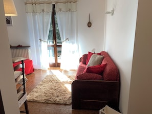 A cozy living room with a red sofa, a wooden chair, a fluffy rug, and a view through a window with sheer curtains. - AffittiPontediLegno - Sorgente (Ponte di legno)
