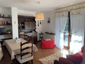 A dining area with a wooden table, chairs, and a red ottoman. There is a kitchen area with cabinets and a hanging light fixture. - AffittiPontediLegno - Sorgente (Ponte di legno)