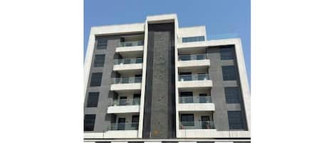 A multi-story building with balconies, a central vertical feature, and a parking area with a truck.