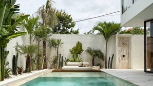 Una piscina con zona de estar integrada, rodeada de plantas tropicales y un edificio moderno.