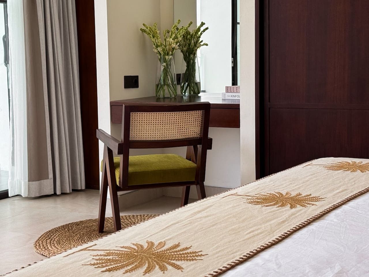 Een kamer met een bed, een houten stoel met een groene kussens, een tafel met vazen met bloemen, en een donkerhouten deur.