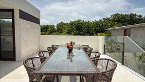 Un área de comedor al aire libre con una mesa de vidrio y sillas de mimbre, rodeada por un balcón con vistas a zonas verdes.