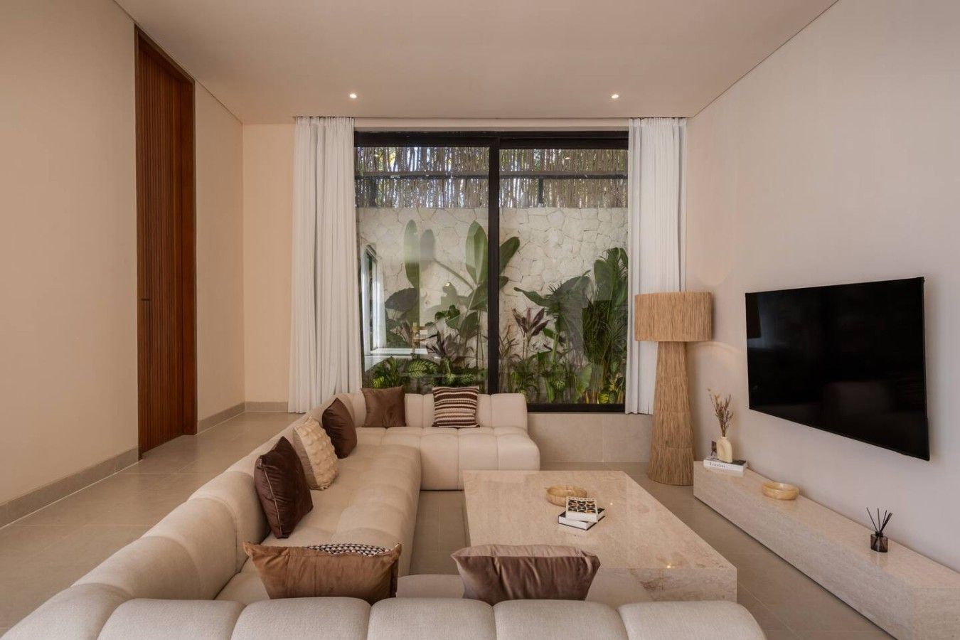 A modern living room with a sectional sofa, a flat-screen TV, and a large window with a view of greenery.