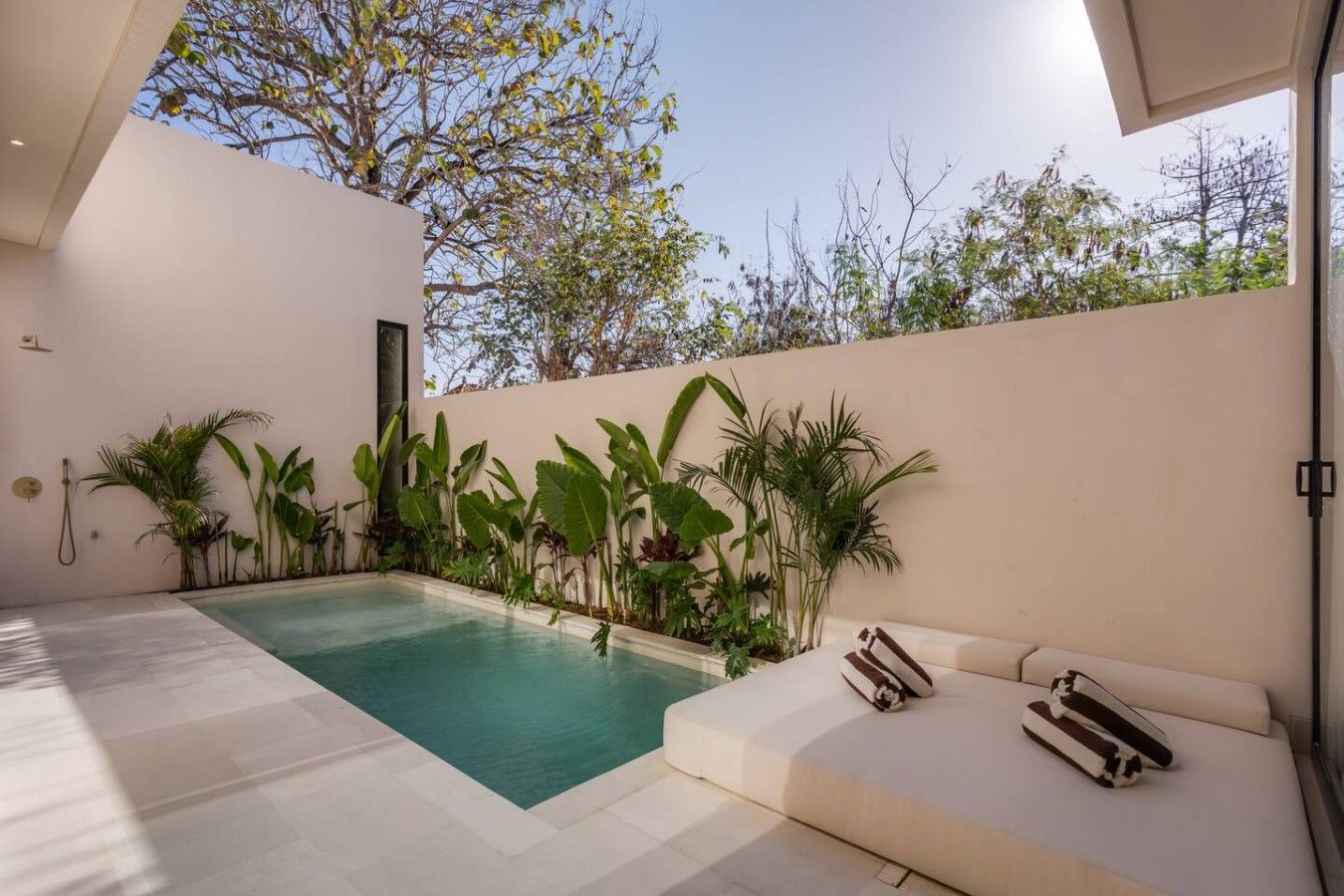 A modern outdoor pool area with a white tiled floor, a beige wall, and a variety of tropical plants.