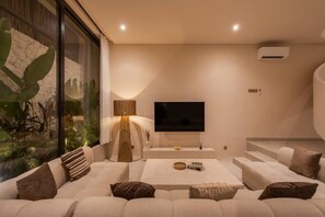 A modern living room with a large sofa, a flat-screen TV, and a large window with plants. - NEW Stylish 2BR Villa w/ Pool & Rooftop, Bingin (South Kuta)