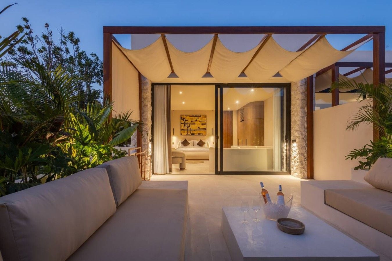 A modern outdoor seating area with a sofa and a table, leading to a glass door entrance to a bedroom with a bed and a bathtub.