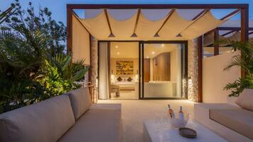 A modern outdoor seating area with a sofa and a table, leading to a glass door entrance to a bedroom with a bed and a bathtub.