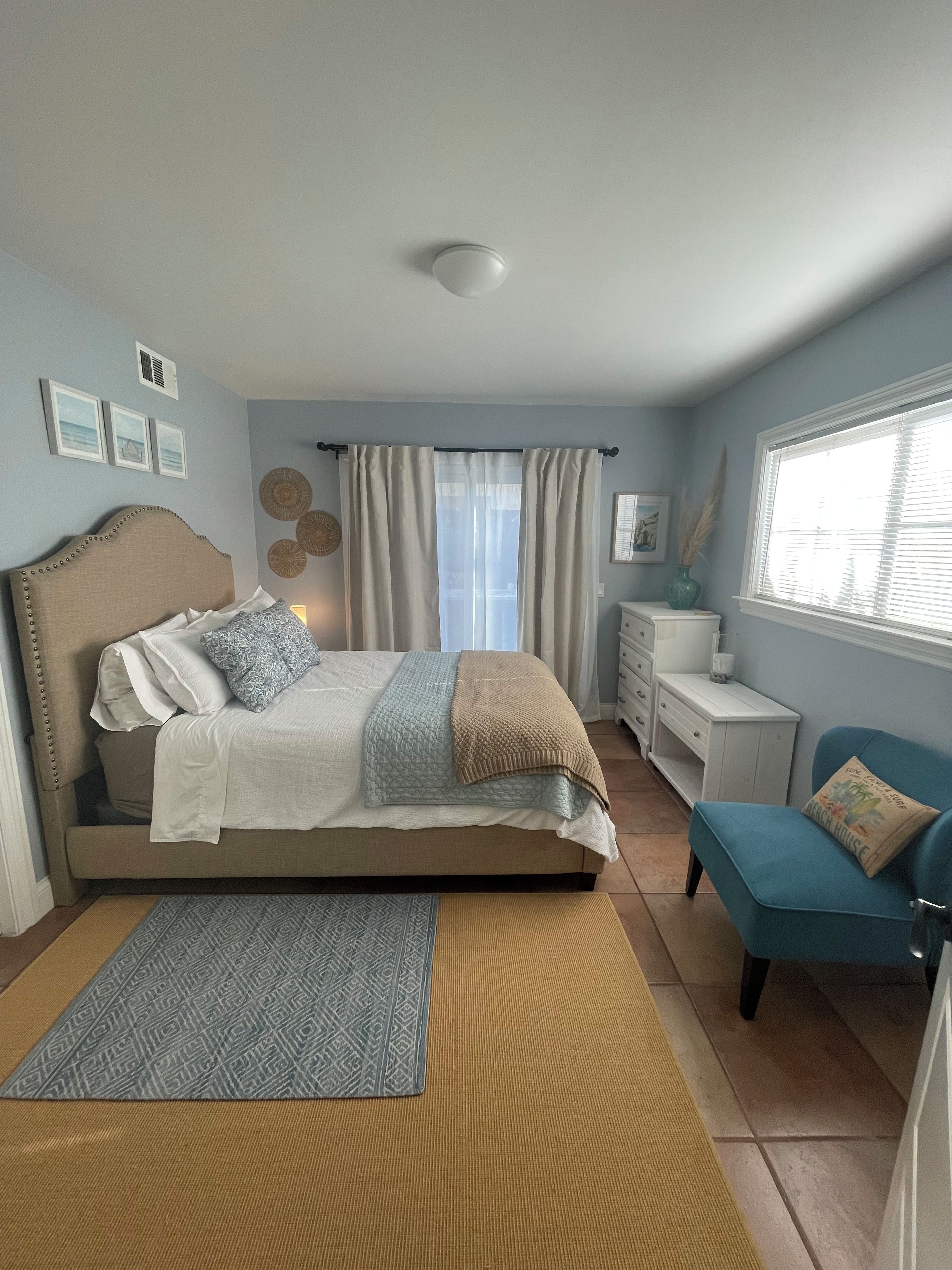 Ein Schlafzimmer mit einem Bett, Nachttischen, einem Fenster mit Vorhängen und einem blauen Sessel.