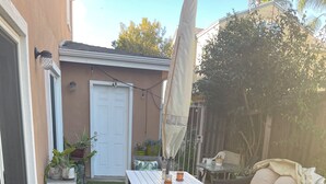 Een achtertuin met kunstgras, een witte tafel, een houten stoel, een parasol en potplanten.