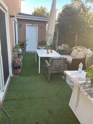 Un patio con césped artificial, una mesa blanca, una silla de madera, una sombrilla y plantas en macetas.