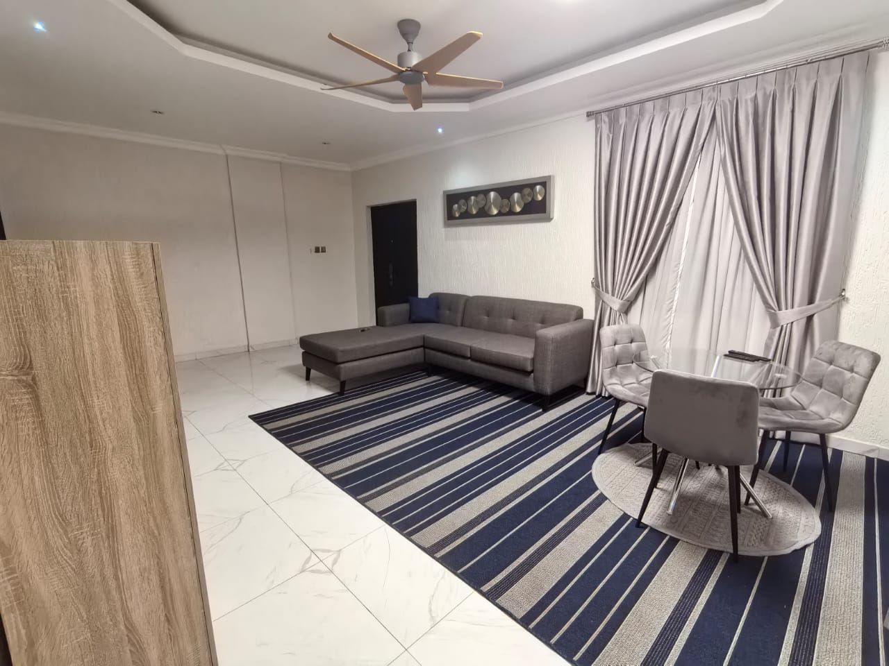 A modern living room with a sectional sofa, a dining table with chairs, a ceiling fan, and a large window with curtains.
