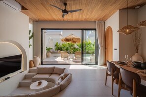 A modern living room with a curved sofa, a dining area, and a view of a tropical garden through glass doors. - Orion 2BR Private Pool, Sauna & Ice Bath in Bingin (South Kuta)