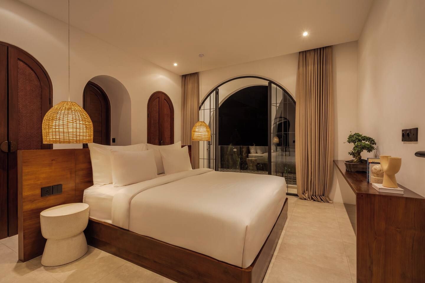 A modern bedroom with a large bed, two bedside lamps, a wooden headboard, a round bedside table, and a view through a glass door.