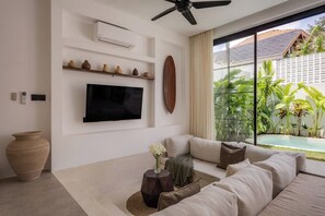 Een moderne woonkamer met een flatscreen televisie, een plafondventilator en een groot raam met uitzicht op een zwembad en groen.