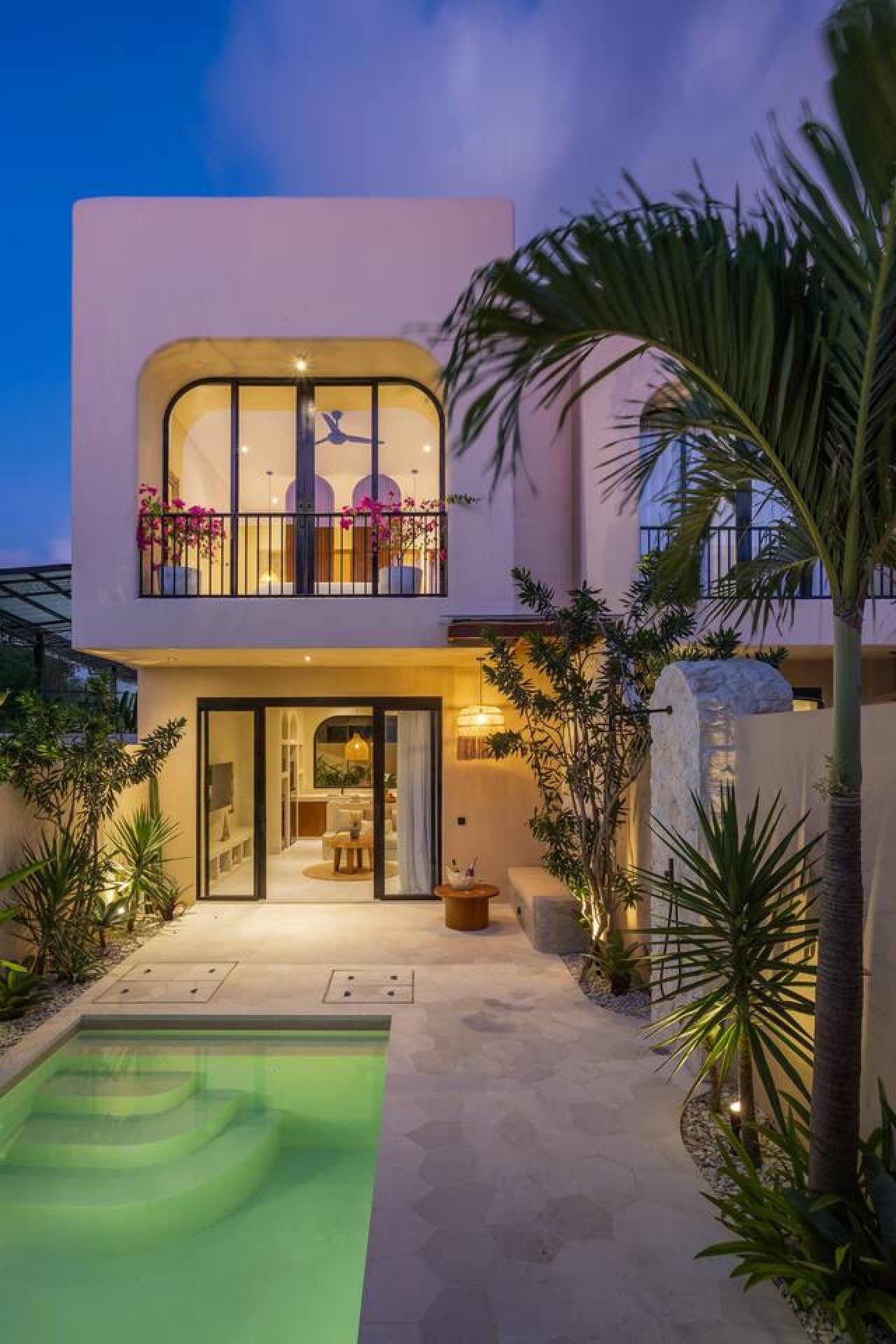 A modern two-story house with a balcony, a swimming pool, and tropical plants.