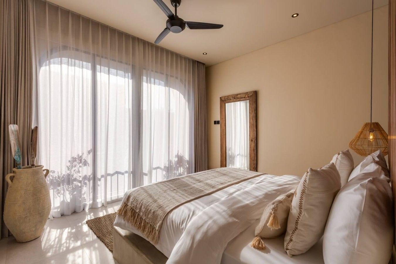 Quarto com cama, ventilador de teto, uma janela grande com cortinas leves e um espelho com moldura de madeira.
