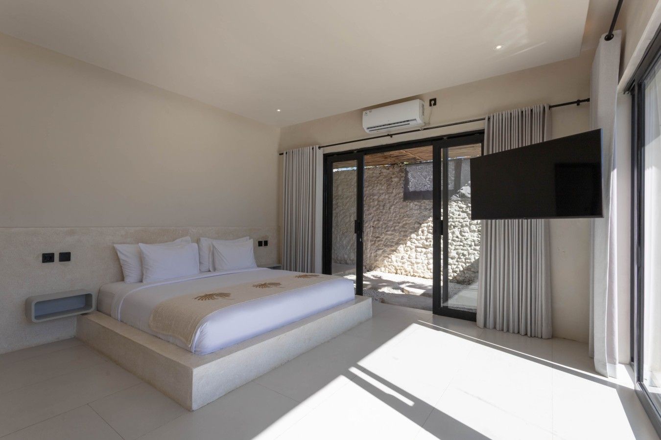 A modern bedroom with a large bed, a flat-screen TV, and a sliding glass door leading to a stone exterior.