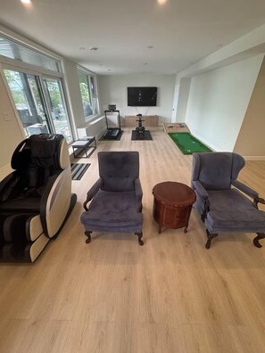 A spacious living room with a massage chair, two armchairs, a small round table, and exercise equipment.