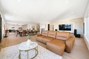 Un vaste espace de vie comprenant un canapé en cuir, une table à manger avec des chaises, une cuisine à l’arrière-plan et un grand téléviseur à écran plat fixé au mur.