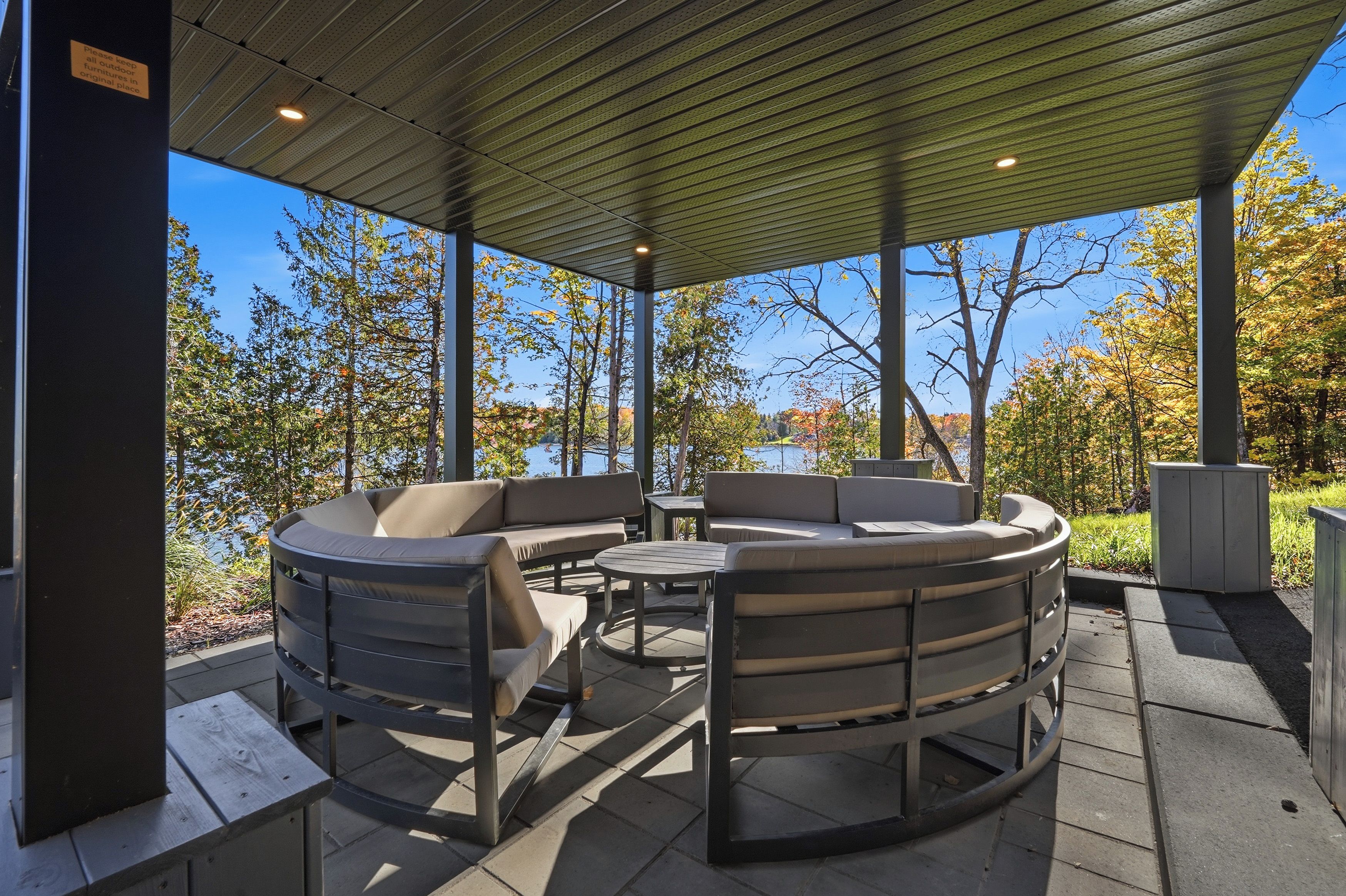An outdoor patio with a round table and chairs, a covered area with a metal roof, and a view of trees and a body of water.