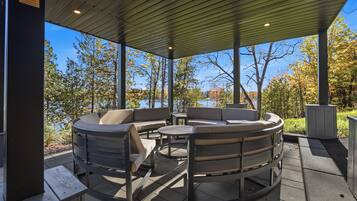 An outdoor patio with a round table and chairs, a covered area with a metal roof, and a view of trees and a body of water.