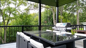 An outdoor patio with a glass-top table and chairs, a grill, and a view of a wooded area.