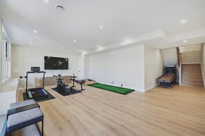 Une pièce spacieuse dotée d’un plancher en bois, d’un tapis de course, de vélos d’appartement, d’un tapis de sol vert pour les exercices et d’un téléviseur fixé au mur.